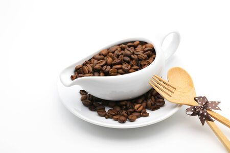 Coffee beans in a ceramic milk jug with fork and spoon on white backgroundの写真素材