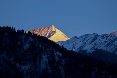 sunrise on the top of a snow-covered mountain. The mountain in the front is covered with pine trees.の写真素材