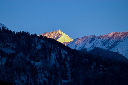 sunrise on the top of a snow-covered mountain. The mountain in the front is covered with pine trees.の写真素材