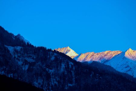 sunrise on the top of a snow-covered mountain. The mountain in the front is covered with pine trees.の写真素材