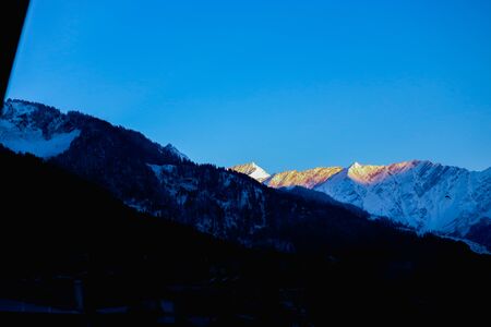 sunrise on the top of a snow-covered mountain. The mountain in the front is covered with pine trees.の写真素材