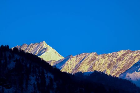 sunrise on the top of a snow-covered mountain. The mountain in the front is covered with pine trees.の写真素材