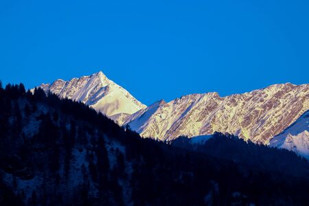 sunrise on the top of a snow-covered mountain. The mountain in the front is covered with pine trees.の写真素材
