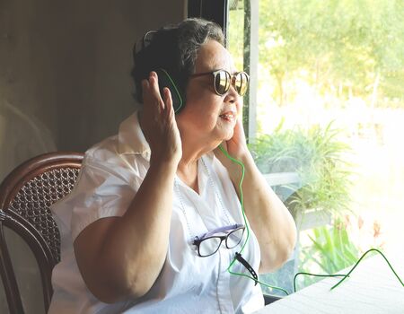 Happy senior woman wearing sunglasses sitting by the window , enjoy listening music from headphone.の写真素材