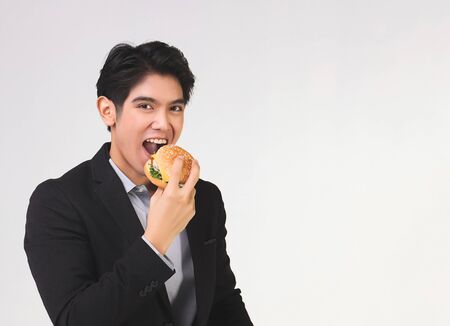 Stylish Asian young and handsome business man wearing dark suit , eating hamburger and looking at camera on white background.の写真素材