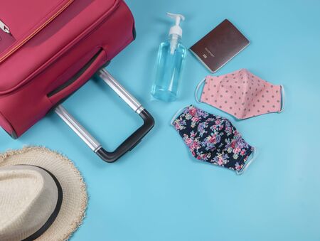 COVID-19 prevention while traveling and new normal lifestyle concept. Top view of two colorful fabric face masks, alcohol hand sanitizer gel ,hat , suitcase and passport on blue background.の写真素材