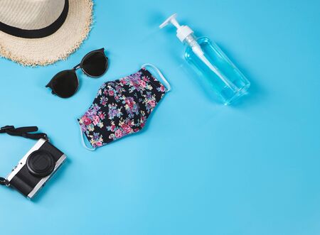 COVID-19 prevention while traveling and new normal lifestyle concept. Top view of flower pattern  fabric face masks, alcohol hand sanitizer gel ,hat and  camera on blue background with copy space.の写真素材
