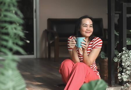 Happy Asian woman wearing red stripe t-shirt sitting in balcony wiht house plants decoration ,holding cup of coffee .smiling happily in morning sunlight.Morning routine,stay home concept.の写真素材