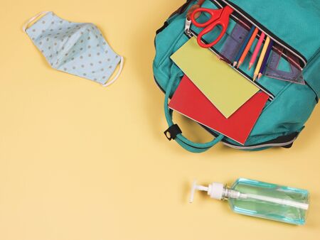 COVID-19 prevention while going back  to school  and new normal  concept. Top view of backpack with school supplies , blue polka dot fabric mask and alcohol sanitizer gel on yellow background with copy space.の写真素材