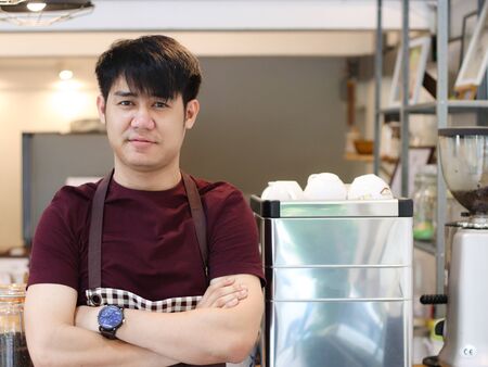 Asian young man standing in front of coffee machine in his own coffee shop , crossing his arms and looking at the camera.の写真素材