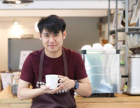Asian young man sitting in front of coffee machine in his own coffee shop , holding mug of coffee and smiling to the camera.の写真素材