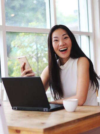 Beautiful Asian woman with long black hair got good news on her mobile phone and smile happily  , surprisingly to the camera. computer and cup of coffee on table.の写真素材
