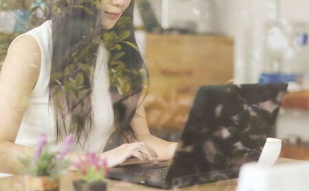 Cropped image through glass window of  beautiful Asian business woman with long black hair working in cafe , typing on computer answer  email with her customer.の写真素材