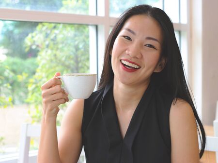 Beautiful Asian woman with long black hair sitting in cafe holding cup of coffee laughing and...