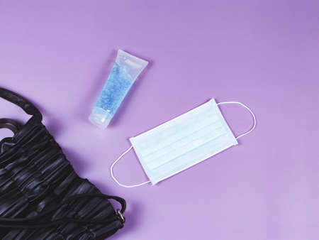 Top view or flat lay of woman's handbag with  surgical hygienic face  mask and alcohol sanitizer gel on purple background with copyspace. Protection from COVID-19 and new normal lifestyle  concept.の写真素材