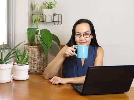 Asian woman wearing eye glasses sitting at wooden table wiht computer laptop and house plant indoor, drinking coffee with blue cup, smiling and looking at camera. working from home concept.の写真素材