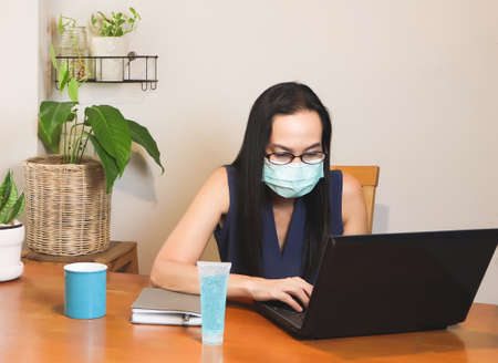 Asian woman wearing hygienic mask sitting at wooden table indoor with computer notebook,alcohol sanitizer gel,typing on computer. working from home.protection from covid-19 concept.の写真素材