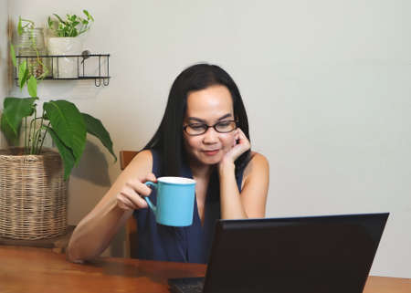 Asian woman sitting at wooden table indoor , holding cup of coffee and looking at computer screen on the table.Working from home concept.の写真素材