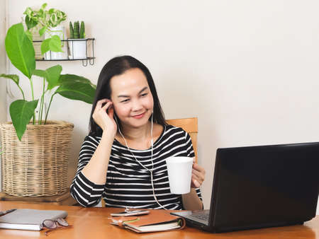 Asian woman sitting at wooden table indoor with computer, listening music happily from mobile phone, relaxing while working at home concept.の写真素材