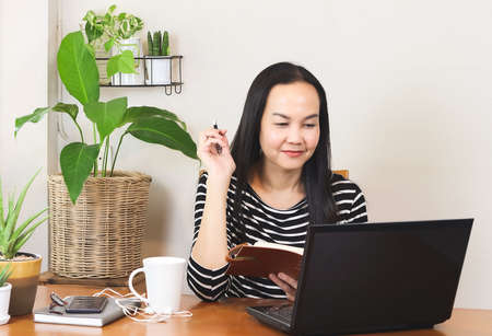 Asian woman sitting at wooden table decorated with houseplant , video chatting with laptop and holding notebook and pen in her hand .remote working or  working from home concept.の写真素材