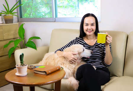 Asian woman sitting on couch in living room  with her golden retriever dog holding cup of coffee smile and looking at camera.selective focus on woman.の写真素材