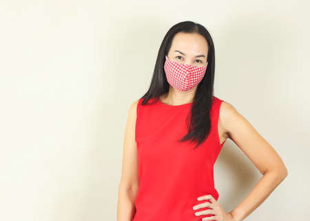 Beautiful Asian woman wearing red and white checkered hygienic mask and red no sleeve t-shirt,standing on white background , hand on her waist   and looking at camera.Fashion and protection from covid-19 concept.の写真素材