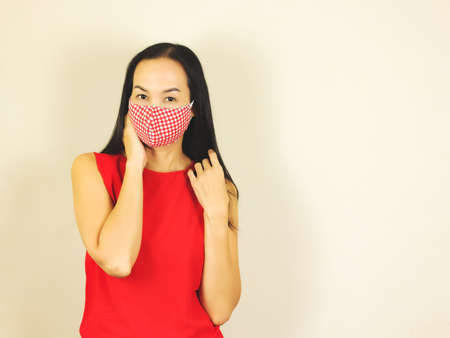 Beautiful Asian woman wearing red and white checkered hygienic mask and red no sleeve t-shirt,touching her cheek and hair  and looking at camera.Fashion and protection from covid-19 concept.の写真素材