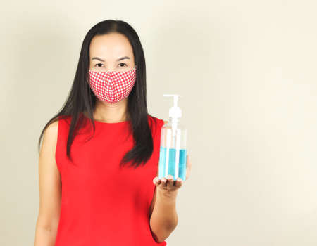 Beautiful Asian woman wearing red and white checkered hygienic mask and red no sleeve t-shirt,looking at camera , holding alcohol sanitizer hand gel.Fashion and protection from covid-19 concept.の写真素材