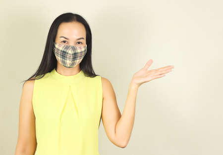 Beautiful Asian woman wearing yellow tartan hygienic mask and yellow no sleeve t-shirt,showing copy space by her hand..Fashion and protection from covid-19 concept.の写真素材