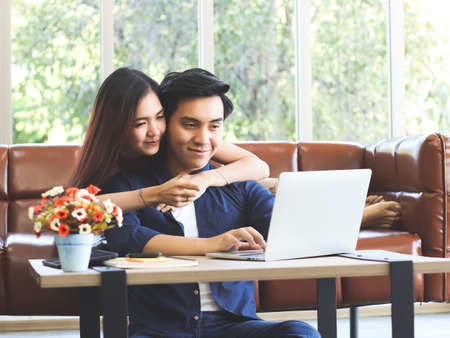 Attractive Young Asian couple sitting close together  on couch in living room with computer laptop on woman's lap , a man hugging her and woman looking at camera with finger on lips.の写真素材