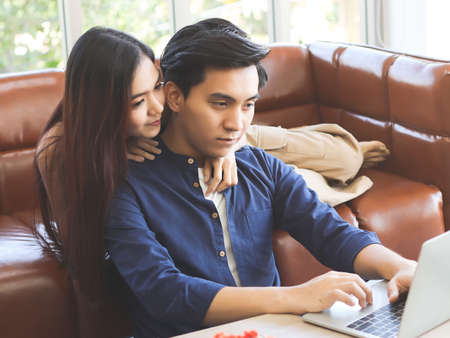 Young Asian couple sitting on couch in living room, a man playing game on smart phone while woman pouting to him.の写真素材