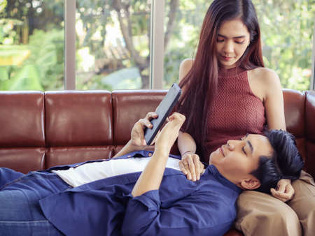 Young Asian couple sitting close together on couch in living room , looking  happily at camera  while woman open the gift box which  a man gave her.の写真素材