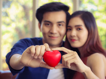 Young Asian couple sitting close together on couch in living room , looking  happily at camera ...