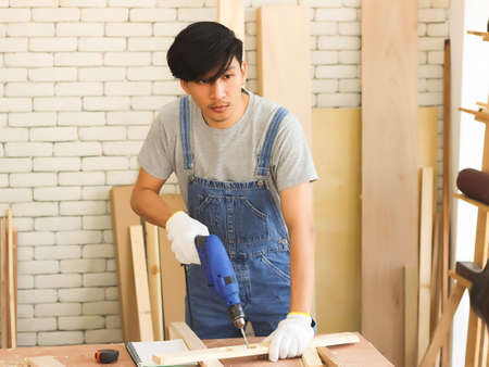 Young and handsome Asian repairman working in workshop with white brick background, drilling wood...