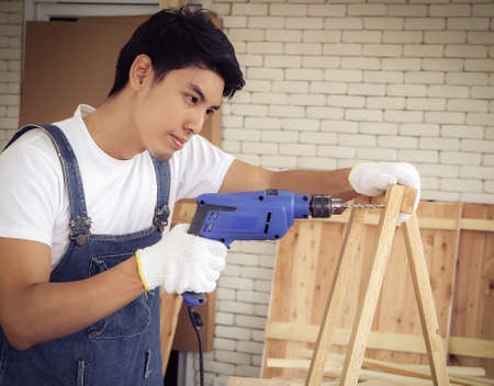 Handsome young Asian repairman wearing yellow safety helmet ,working in wood workshop with white brick background , using grinding machine blur foreground of denim jeans.の写真素材