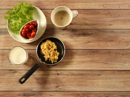 Top view of breakfast set , scramble eggs in pan , vegetable and tomato , cup of coffee and  milk  on wooden background.の写真素材
