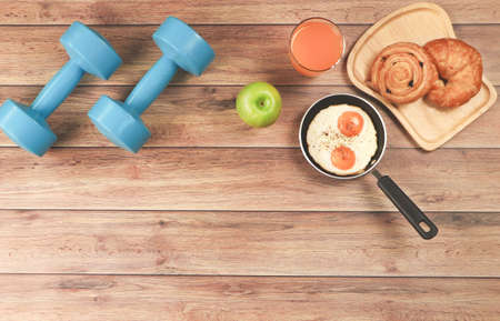 Top view of blue dumbbells , apple ,orange juice, fried eggs and pastry with copy space on wooden background. Healthy lifestyle concept healthy breakfast and excercise.の写真素材