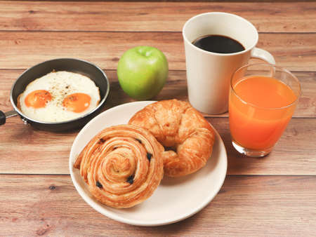 Front view of breakfas set , danish pastry and croissant, fried eggs in pan, green apple, black coffe and orange juice on wooden table.Healthy breakfas concept.の写真素材