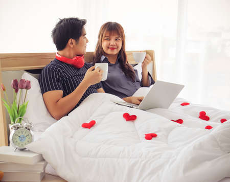 Young and attractive sitting on bedroom holding cups of coffee looking to each other with love , many red hearts on blanket,Valentine's day concept.の写真素材