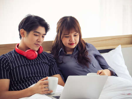 Young and attractive couple  sitting on bed holding cups of coffee looking to screen of computer laptop ,Valentine's day concept.の写真素材