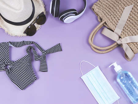 COVID-19 prevention , travel and new normal concept, top view of   woven bag with  sanitizer gel  and surgical  face masks  with woman's vacation accessories on purple  background with copy space.の写真素材