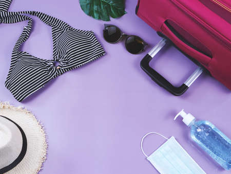 COVID-19 prevention , summer and new normal concept, top view of   bikini swimsuit and women's vacation accessories with surgical mask and sanitizer gel  on purple backgroundの写真素材