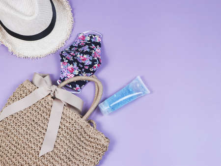 COVID-19 prevention , travel and new normal concept, top view of   woven bag with  sanitizer gel  , flowery pattern face mask and hat  on purple  background with copy space.の写真素材