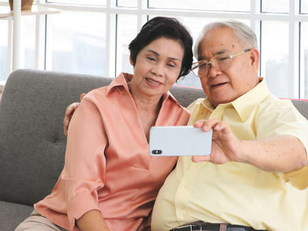 Happy Asian senior couple having good time together,sitting in living room , talking selfie photo or video chatting looking at  smart phone. Elderly lifestyle and technology concept.の写真素材