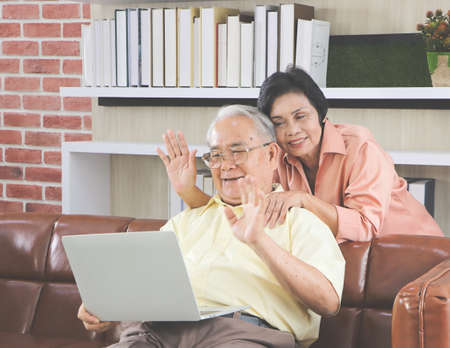 Senior people lifestyle and technology  concept. Happy Asian elderly couple making video call on laptop, Senior man sitting on sofa while his wife hugging him from behind waving on computer screen and smiling..の写真素材
