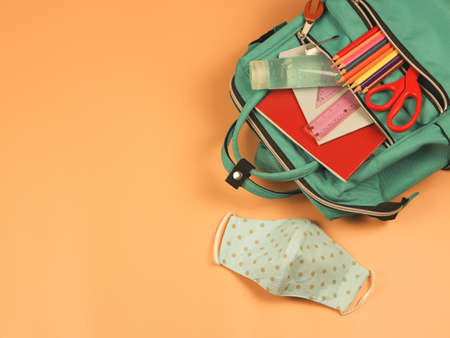 Top view or flat lay of backpack with school supplies ,blue  polka dot fabric masks and sanitizer gel on orange background. COVID-19 prevention while going back  to school  and new normal  conceptの写真素材