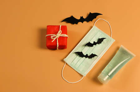 Top view or flat lay of red gift box with black paper bats , medical face mask and alcohol sanitizer gel  on orange background with copy space. Halloween present concept , Covid19 protection and new normal concept.の写真素材