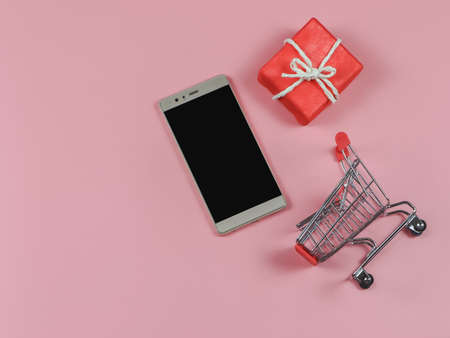 Top view or flat lay of mobile phone red gift box and shopping cart on pink background with copy space. Christmas holiday and online shopping concept.の写真素材