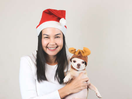 Portrait of Asian woman wearing  red Santa Christmas hat  smiling and looking at camera, holding  her Chihuahua dog wearing reindeer horn hat making funny face. Christmas concept.の写真素材