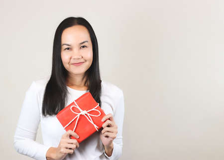 Portrait of pretty Asian woman in white long sleeve  t-shirt holding red gift box, smiling and  looking at camera, front view on white background with copy space.の写真素材